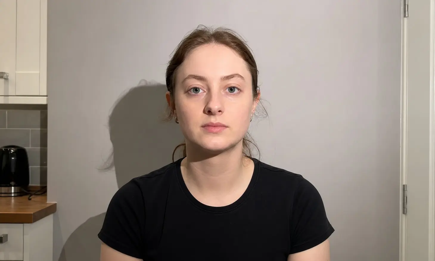 UK-style passport portrait taken in a kitchen with a harsh shadow under the chin and a dark halo on the wall behind the head